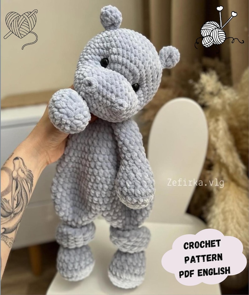 Puede incluir: Un juguete de peluche de hipop&oacute;tamo gris tejido a crochet con una superficie texturizada. El juguete tiene una nariz en forma de coraz&oacute;n y peque&ntilde;os ojos negros. El juguete est&aacute; siendo sostenido por una mano con un tatuaje.