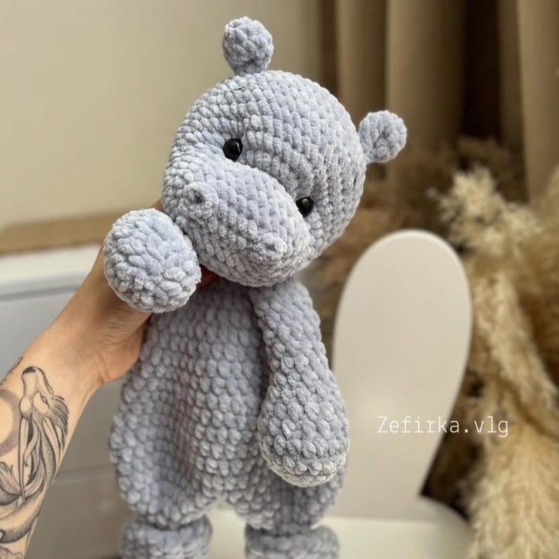Puede incluir: Un juguete de peluche de crochet suave de color gris con forma de hipop&oacute;tamo. El juguete tiene una superficie texturizada y peluda y ojos negros. El juguete est&aacute; siendo sostenido por una mano.