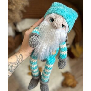 May include: A handmade crochet gnome doll with a light blue hat and striped blue and white outfit. The gnome has a long white beard, gray gloves, and gray shoes. A gold-colored charm hangs from the gnome's chest.