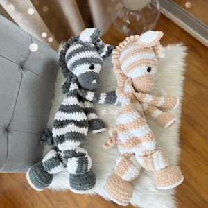 May include: Two handmade crochet zebra stuffed animals. One is gray and white striped, the other is peach and white striped. The text "juk.toys" is visible on the left side. The zebras are lying on a white fluffy surface.
