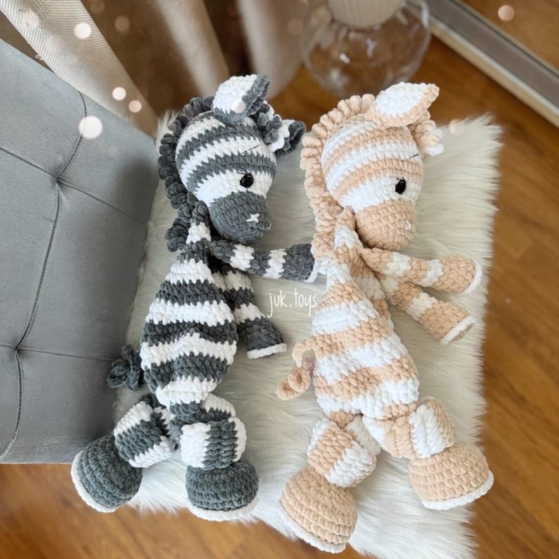 May include: Two handmade crochet zebra stuffed animals. One is gray and white striped, the other is peach and white striped. The text "juk.toys" is visible on the left side. The zebras are lying on a white fluffy surface.