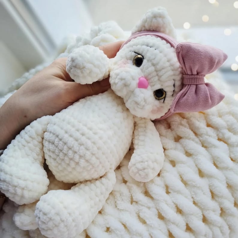 Puede incluir: Mu&ntilde;eca de gato de ganchillo blanco hecha a mano con lazo y diadema rosas. El gato tiene ojos negros, mejillas rosas y nariz rosa. La mu&ntilde;eca est&aacute; sobre una manta blanca texturizada. Una mano sostiene la mu&ntilde;eca.