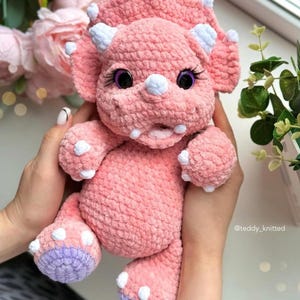 May include: A pink crocheted triceratops stuffed animal with white horns and purple feet. The toy has large black eyes with white eyelashes and a pink open mouth.