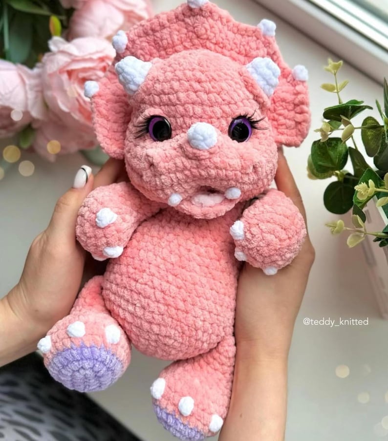 May include: A pink crocheted triceratops stuffed animal with white horns and purple feet. The toy has large black eyes with white eyelashes and a pink open mouth.