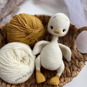 May include: A white crocheted goose toy with yellow feet and a black beak. The goose is sitting in a wicker basket with two balls of yarn, one white and one yellow.