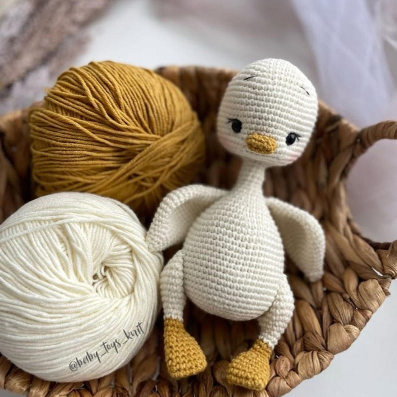 May include: A white crocheted goose toy with yellow feet and a black beak. The goose is sitting in a wicker basket with two balls of yarn, one white and one yellow.