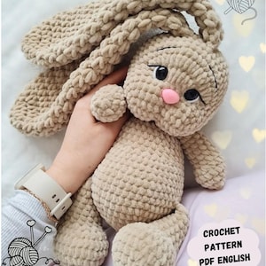 Puede incluir: Un juguete de peluche de conejo tejido a crochet de color marrón claro. El conejo tiene orejas largas, una nariz rosa y ojos negros. El juguete está siendo sostenido por una mano. La imagen incluye el texto "CROCHET PATTERN PDF ENGLISH".