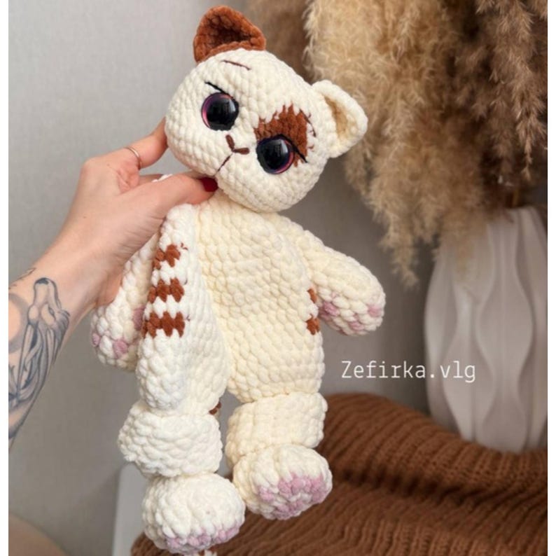 May include: A handmade crochet cat plush toy in cream with brown and pink accents. The cat has large, expressive eyes and a stitched face. The text "Zefirka.vlg" is visible on the right side of the image.