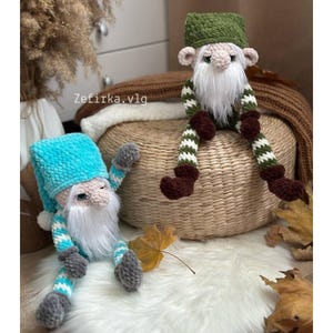 May include: Two handmade crochet gnomes with long white beards and hats. One gnome has a blue hat and striped legs, the other has a green hat and green and white striped legs. The gnomes are sitting on a woven basket and a white fluffy surface.