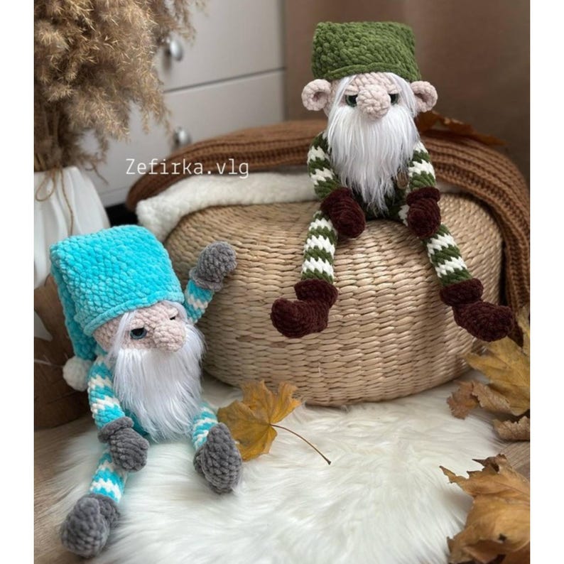 May include: Two handmade crochet gnomes with long white beards and hats. One gnome has a blue hat and striped legs, the other has a green hat and green and white striped legs. The gnomes are sitting on a woven basket and a white fluffy surface.