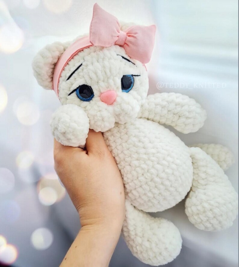 Puede incluir: Un juguete de peluche de gato blanco hecho a mano con ojos azules, una nariz rosa y una diadema con lazo rosa. El gato est&aacute; tejido con un hilo suave y texturizado. El juguete se sostiene en una mano.