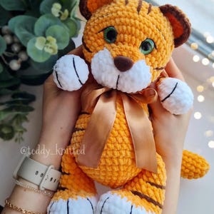 May include: A handmade orange and brown crochet tiger plush toy with a brown ribbon. The tiger has white paws and a white muzzle. The tiger's eyes are green. The text "@teddy_knitted" is visible on the image.