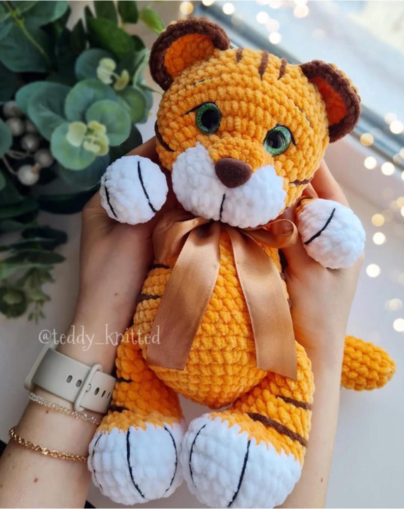 May include: A handmade orange and brown crochet tiger plush toy with a brown ribbon. The tiger has white paws and a white muzzle. The tiger's eyes are green. The text "@teddy_knitted" is visible on the image.