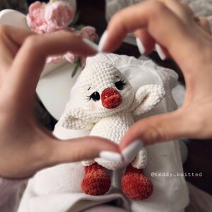 May include: A handmade white crochet duckling with a red beak and feet. The duckling has long eyelashes and is posed with its arms outstretched. The image is framed by hands forming a heart shape.