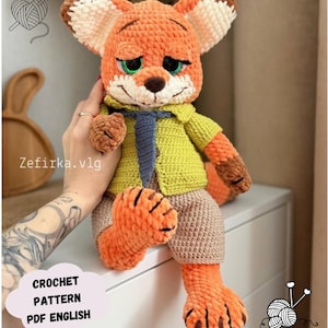 Puede incluir: Muñeco de zorro tejido a crochet, con pelaje naranja, orejas color crema y ojos verdes. El zorro viste una camisa amarilla, una corbata azul y pantalones cortos color beige. Incluye el texto "CROCHET PATTERN PDF ENGLISH".