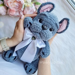 May include: A crocheted plush toy in the shape of a French bulldog. The toy is blue with pink ears and a white bow. The toy is being held by a person's hand. The toy is made with soft yarn and has a sweet expression.