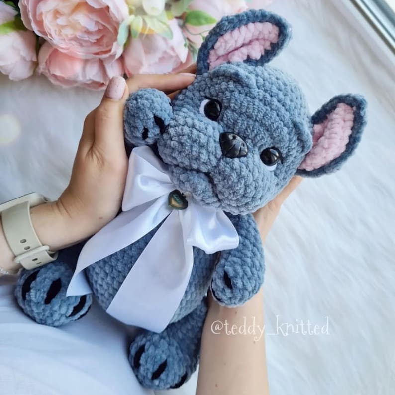 May include: A crocheted plush toy in the shape of a French bulldog. The toy is blue with pink ears and a white bow. The toy is being held by a person's hand. The toy is made with soft yarn and has a sweet expression.