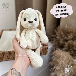 May include: A crocheted white bunny rabbit amigurumi toy with brown eyes and a brown nose. The bunny is being held by a hand with a tattoo. The image includes the text "CROCHET PATTERN PDF ENGLISH".