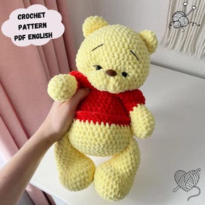 May include: A crocheted bear in yellow and red. The bear has a yellow head, arms, and legs, and a red shirt. The text "CROCHET PATTERN PDF ENGLISH" is in a white cloud-shaped text box. The bear is soft and plush.