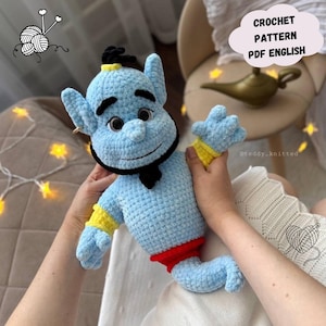 Può includere: Un peluche all'uncinetto azzurro a forma di genio con capelli, sopracciglia e barba neri. Il personaggio indossa polsini gialli e pantaloni rossi. Un fumetto nell'angolo in alto a destra contiene il testo CROCHET PATTERN PDF ENGLISH.