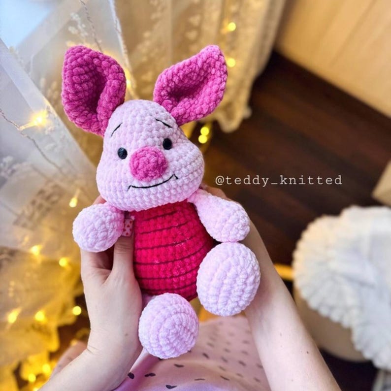 May include: A pink crocheted Piglet plush toy with a red striped shirt. The toy has a pink nose and black eyes. The toy is being held by a person's hands. The Instagram handle @teddy_knitted is visible in the image.