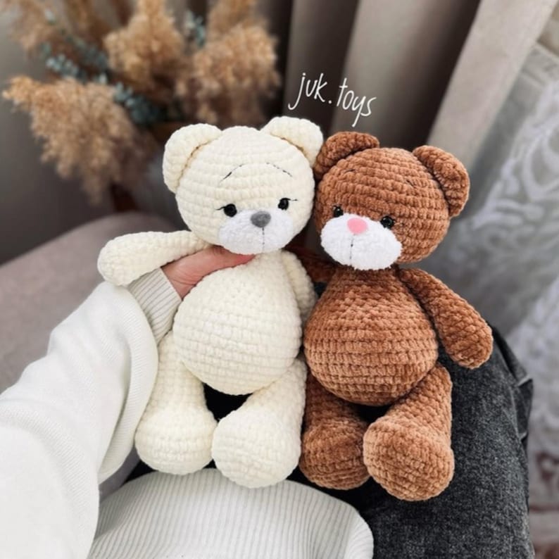 Op de afbeelding: Twee gehaakte teddyberen, een witte en een bruine, zitten samen. De witte beer heeft zwarte ogen en een grijze neus. De bruine beer heeft roze ogen en een bruine neus. Beide beren houden hun poten in de lucht.