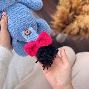 May include: A blue crocheted stuffed animal with a pink bow and black yarn hair. The animal has a brown button on its body.