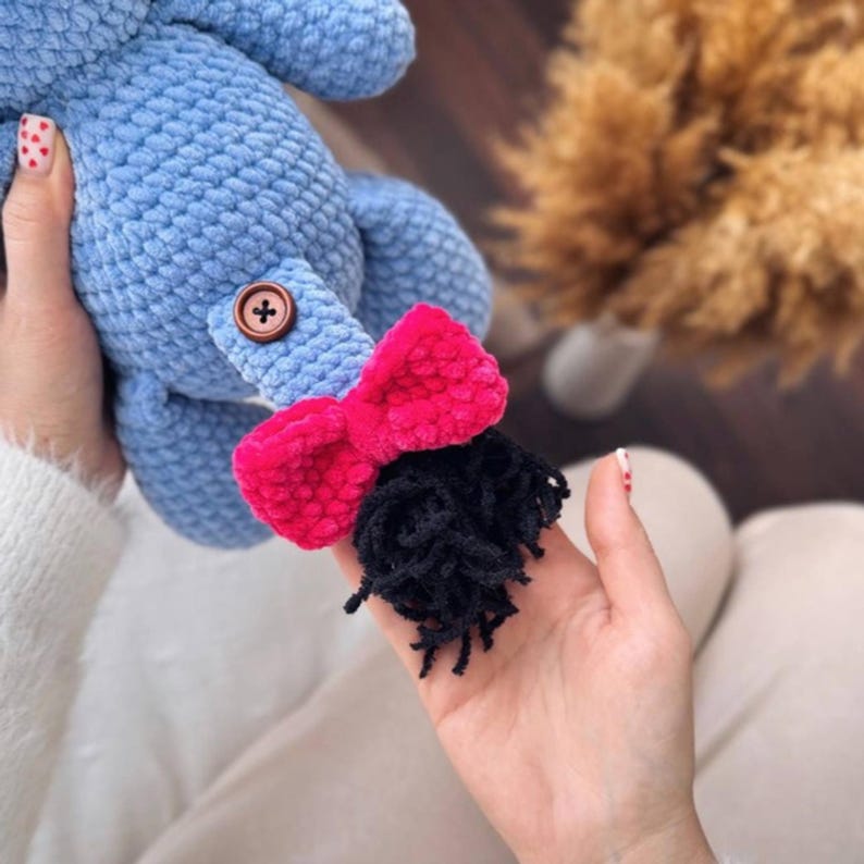 May include: A blue crocheted stuffed animal with a pink bow and black yarn hair. The animal has a brown button on its body.