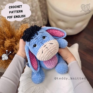 May include: A crocheted blue Eeyore plush toy with pink ears and a black mane. The toy is being held by a person's hands. The text "@teddy_knitted" is visible in the bottom right corner of the image.