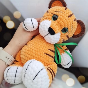 May include: A handmade orange and brown tiger plush toy with white accents. The tiger has green eyes, black stripes, and a green ribbon tied around its neck. The plush toy is soft and cuddly, perfect for children.