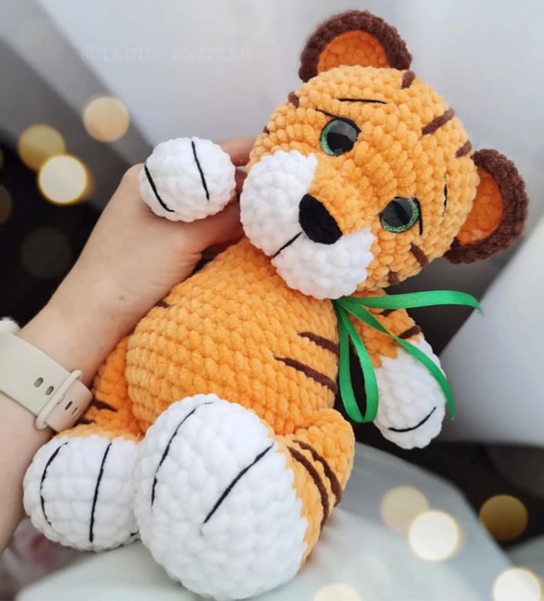 May include: A handmade orange and brown tiger plush toy with white accents. The tiger has green eyes, black stripes, and a green ribbon tied around its neck. The plush toy is soft and cuddly, perfect for children.