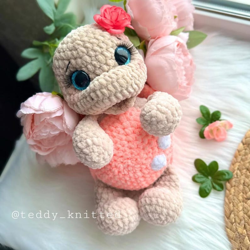May include: A crocheted turtle toy with a pink shell and beige body. The turtle has large blue eyes and a pink flower on its head. The toy is surrounded by pink flowers.
