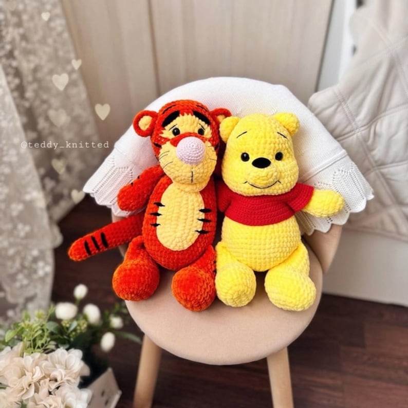 May include: Two crocheted stuffed animals, a yellow and orange tiger and a yellow bear, sitting on a chair. The tiger has a pink nose and black stripes. The bear has a red shirt.