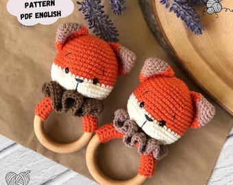 Op de afbeelding: Twee gehaakte vos-rammelaars met houten ringen. De vossen zijn oranje met witte gezichten en bruine accenten. De speelgoedjes liggen op een bruine ondergrond.
