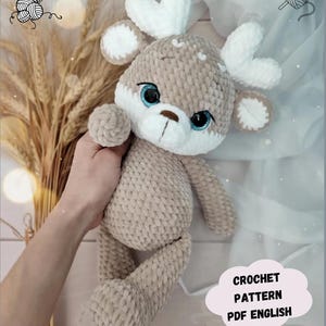 May include: Crocheted plush reindeer toy with white antlers, brown body, and blue eyes. The toy is being held by a hand. The text "CROCHET PATTERN PDF ENGLISH" is visible in the lower right corner of the image.
