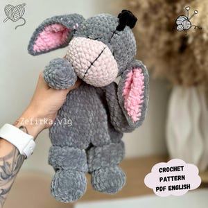Op de afbeelding: Een grijs gehaakt konijn knuffel met roze binnenoren en een roze neus. Het konijn heeft een zwarte vlek op zijn hoofd en is gemaakt van zacht, getextureerd garen. De afbeelding bevat de tekst "CROCHET PATTERN PDF ENGLISH".