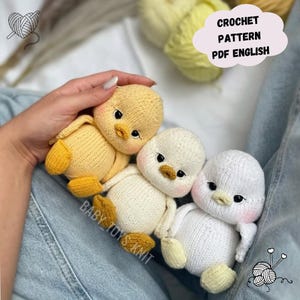 Op de afbeelding: Drie handgemaakte gehaakte eendjes in geel en wit. De eendjes hebben zwarte ogen, oranje snavels en roze wangen. De tekst "CROCHET PATTERN PDF ENGLISH" staat in een wolk-vormige tekstballon. De tekst "BABY TOYS KNIT" is ook zichtbaar.