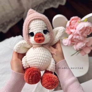 May include: A handmade, white crochet duckling wearing a pink bonnet and featuring orange feet and beak. The duckling has black eyes and long eyelashes. The image is of a soft toy, perfect for a baby or child.