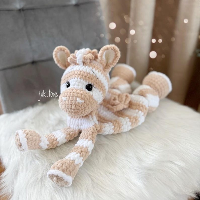 May include: A handmade crochet zebra toy in beige and white stripes. The plush animal has a detailed face with black eyes and is lying down on a white, fluffy surface. The toy is soft and cuddly, perfect for children.