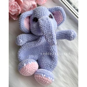 May include: A crocheted purple elephant soft toy with pink ears and feet. The elephant has a textured surface and white stitching around the eyes and mouth.