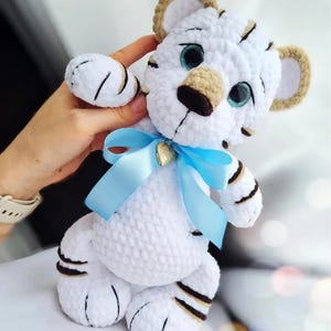 May include: A handmade white crochet tiger cub with brown stripes and accents. The tiger has blue eyes, a tan nose, and ears. A light blue ribbon is tied around its neck, and a small gold heart charm is attached to the ribbon.
