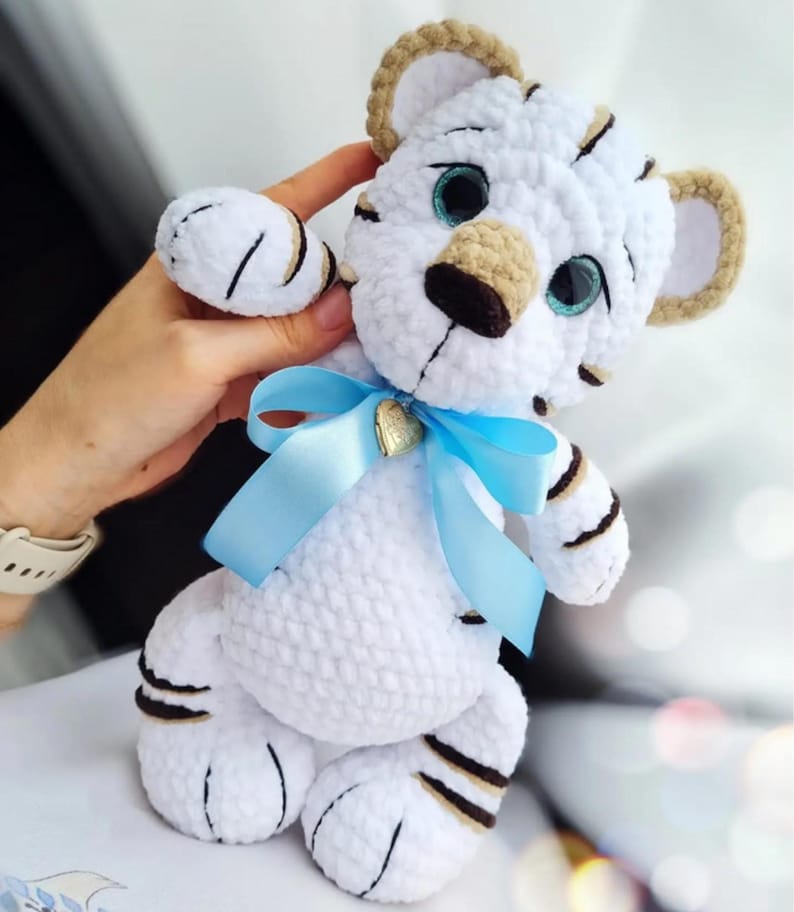 May include: A handmade white crochet tiger cub with brown stripes and accents. The tiger has blue eyes, a tan nose, and ears. A light blue ribbon is tied around its neck, and a small gold heart charm is attached to the ribbon.