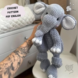 May include: A handmade gray crochet elephant toy with white accents on the ears and feet. The plush animal has a trunk, large ears, and is being held up. The image includes the text "CROCHET PATTERN PDF ENGLISH".