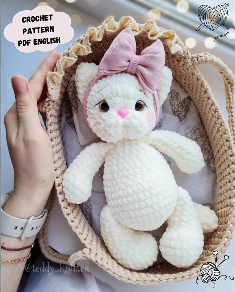 Puede incluir: Mu&ntilde;eca de gato de ganchillo blanco hecha a mano con lazo y diadema rosas, en una cesta de ganchillo beige. La mu&ntilde;eca tiene una nariz rosa y ojos bordados. El texto "CROCHET PATTERN PDF ENGLISH" est&aacute; en una nube.