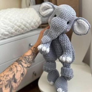 May include: A handmade gray crochet elephant toy with white accents on the ears and feet. The elephant has a trunk, small eyes, and pink cheeks. The toy is soft and plush, perfect for a baby or child.
