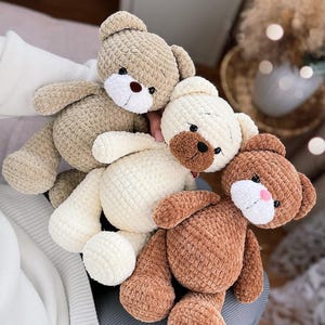 Op de afbeelding: Drie gehaakte teddyberen in verschillende kleuren: bruin, wit en beige. De beren zitten op een grijs oppervlak met de tekst "juk.toys" zichtbaar.