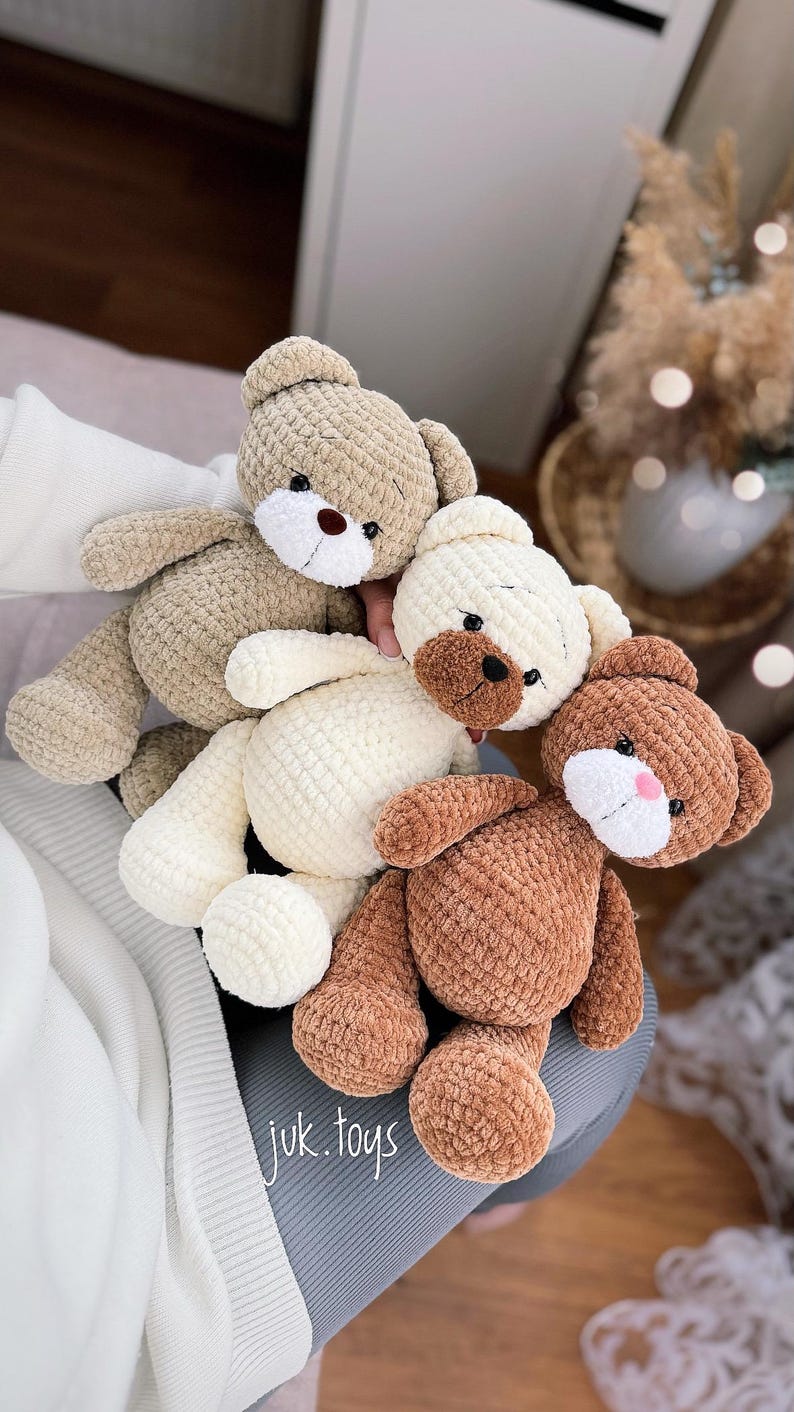 Op de afbeelding: Drie gehaakte teddyberen in verschillende kleuren: bruin, wit en beige. De beren zitten op een grijs oppervlak met de tekst "juk.toys" zichtbaar.