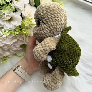 May include: A crocheted plush turtle toy with a green shell and beige body. The turtle has large, black eyes and a white spot on its shell.