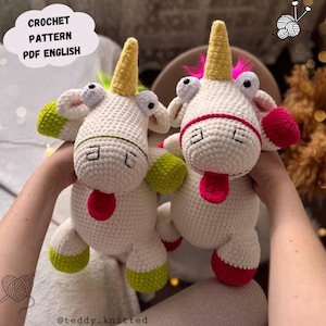 May include: Two white crochet unicorn plushies with yellow horns, green and red accents, and pink tongues. The text "CROCHET PATTERN PDF ENGLISH" is in a cloud-shaped bubble. The unicorns have large, googly eyes and are being held.
