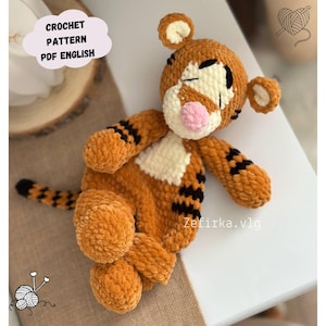May include: A handmade orange and black crochet tiger toy. The tiger has a white muzzle and chest, a pink nose, and black stripes. The image includes text that says "CROCHET PATTERN PDF ENGLISH".